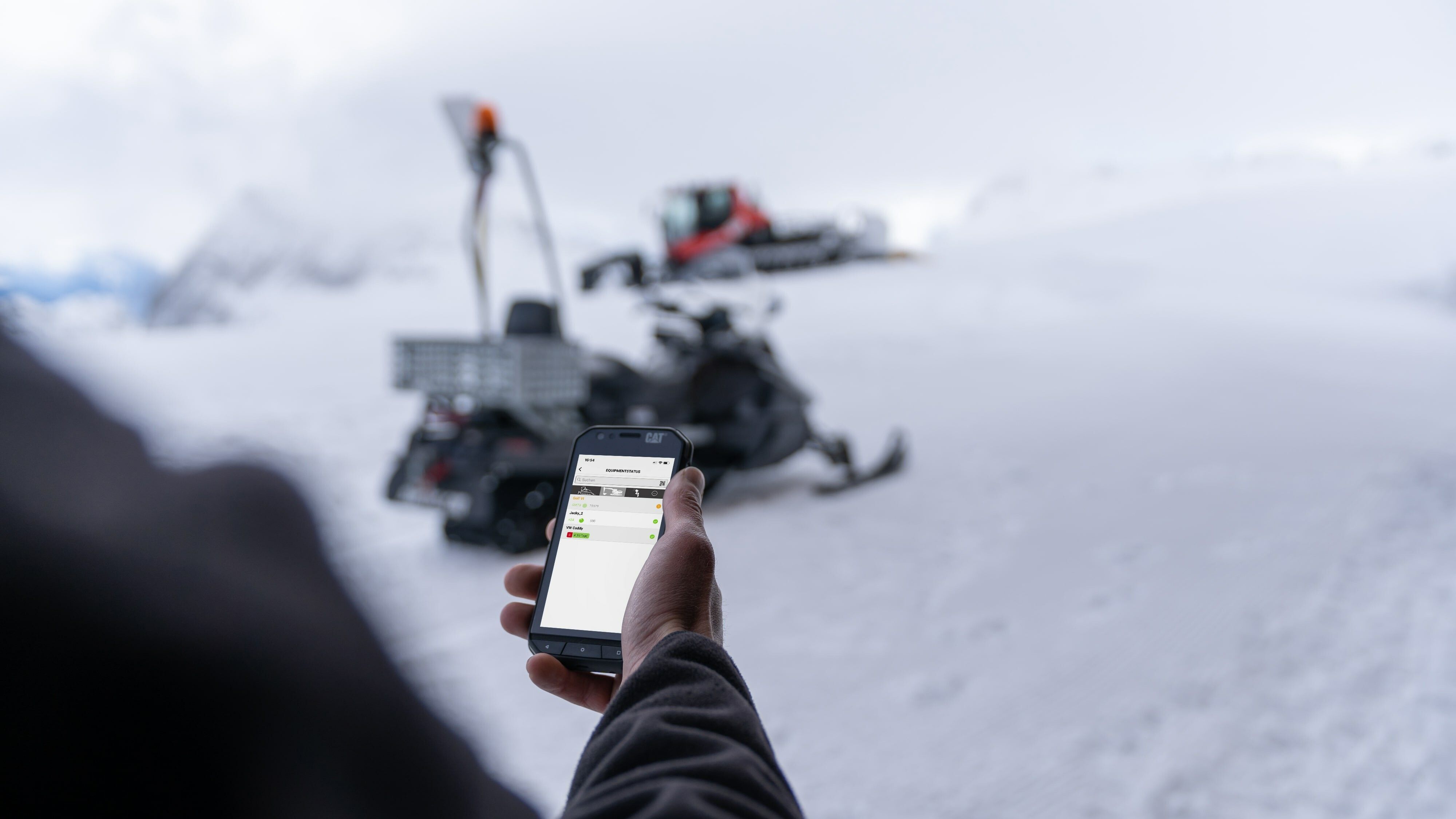 Die SNOWsat App Hand mit Smartphone und SNOWsat App, im Hintergrund Skidoo und PistenBully