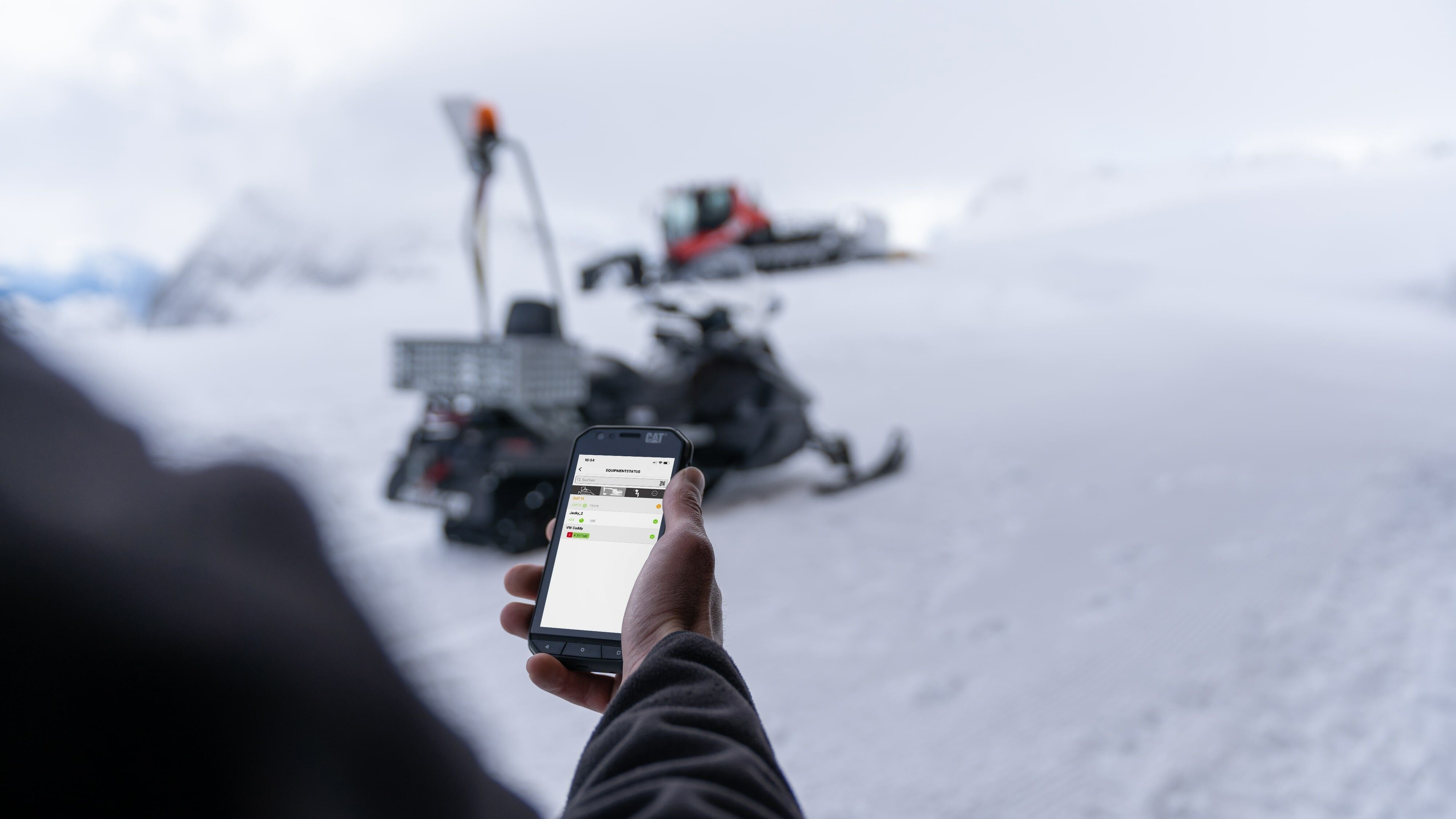 Main avec smartphone et application SNOWsat, en arrière-plan Skidoo et PistenBully